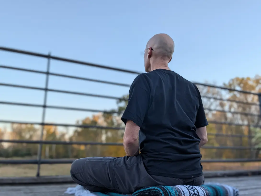 Matthew sitting cross-legged on a folded blanket on The Marsh deck, seen from behind, with autumn trees and blue sky beyond the railing.